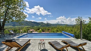 A swimming pool with lounge chairs and a table, surrounded by trees and mountains in the background.