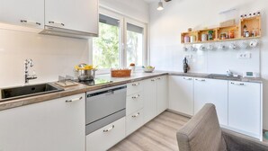A modern kitchen with white cabinets, a built-in oven, and a wooden countertop. There is a window providing natural light and a shelf with kitchen utensils.
