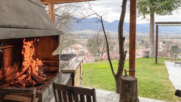 A wood-fired oven with flames, a bench, and a view of a village and mountains.