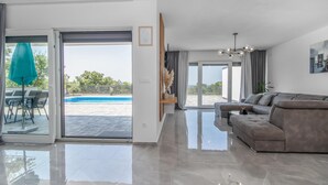 Ein modernes Wohnzimmer mit einem großen grauen Sofa, einem Essbereich mit Tisch und Stühlen sowie einer Glasschiebetür zum Poolbereich.