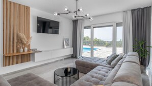 Ein modernes Wohnzimmer mit einem großen grauen Sofa, einem Flachbildfernseher, einer Wand aus Holzlatten und Blick auf einen Pool.