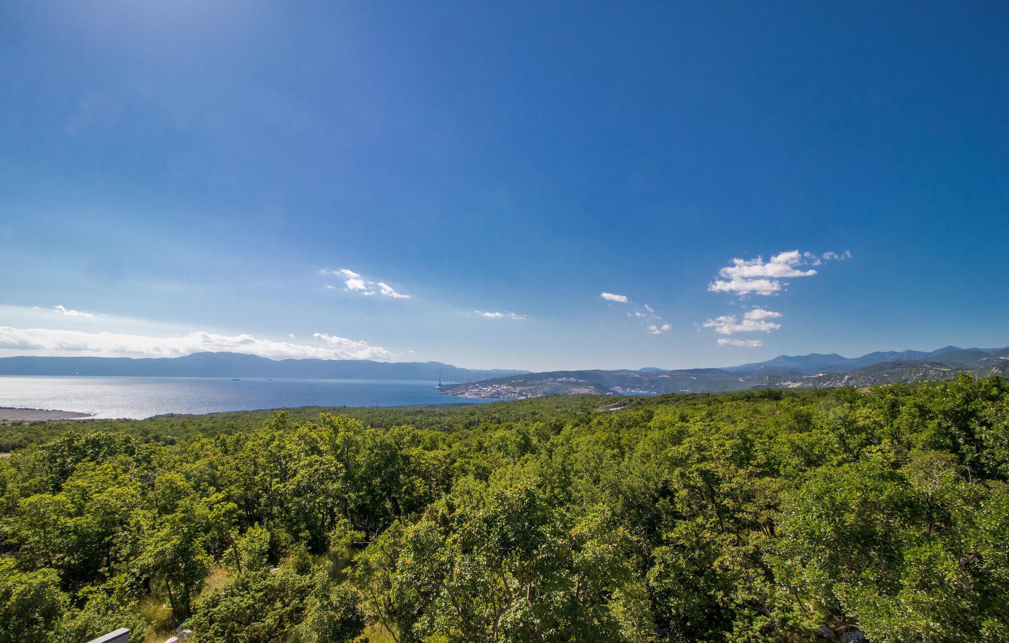 A coastal landscape with dense greenery and a view of the sea and distant mountains.