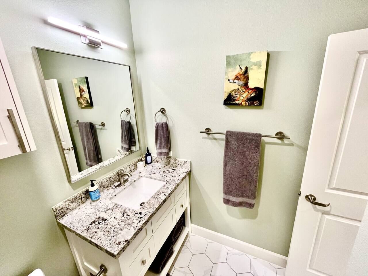 A bathroom with a granite countertop sink, a mirror, a towel rack, and a painting on the wall.