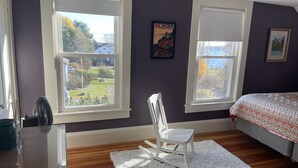 Une chambre avec deux fenêtres, un fauteuil à bascule, un lit recouvert d’une courtepointe à motifs et un tableau encadré accroché au mur.