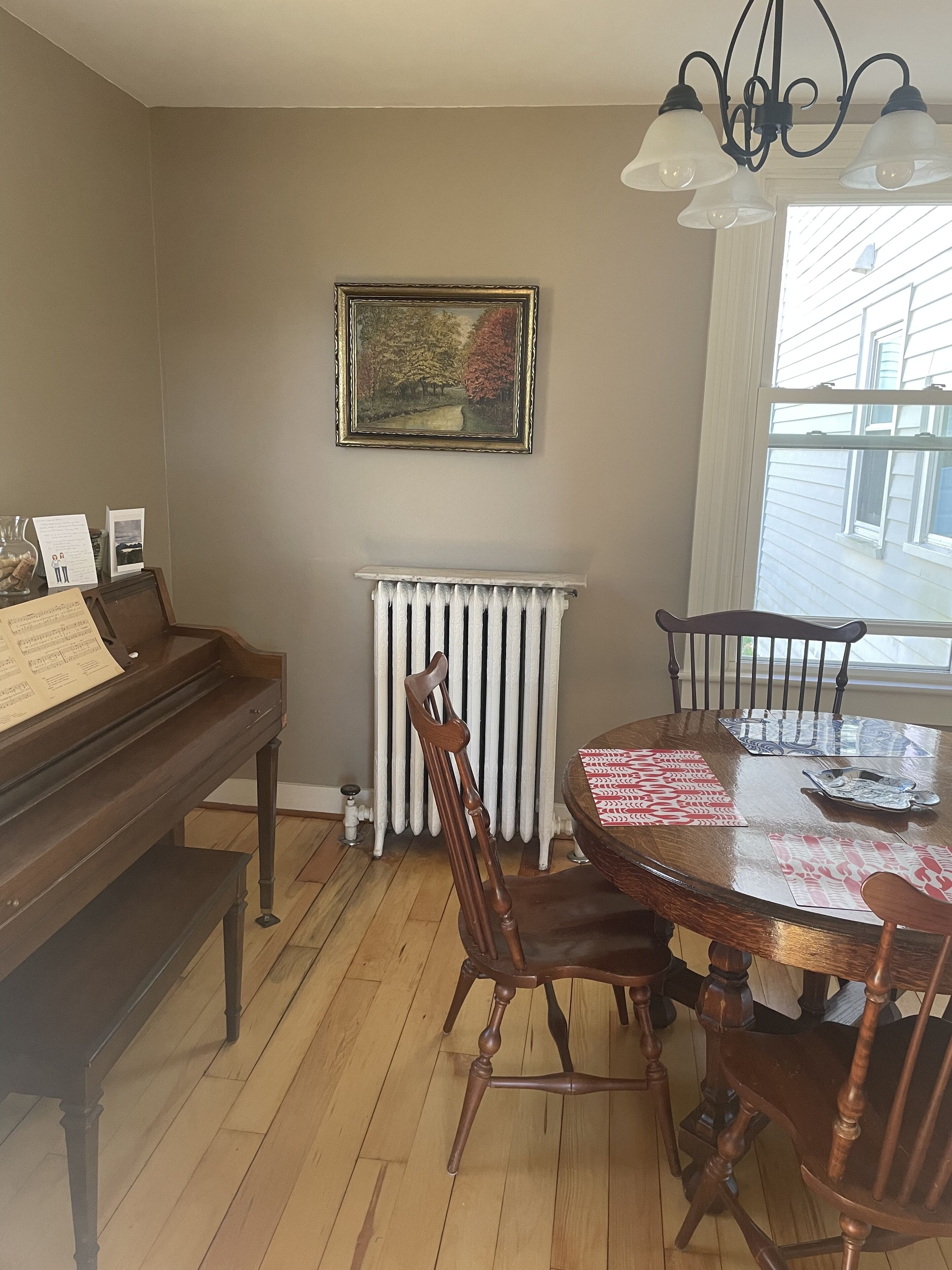 Et spiseområde med et klaver, træstole, et bord med dug, en radiator og et indrammet billede på væggen.