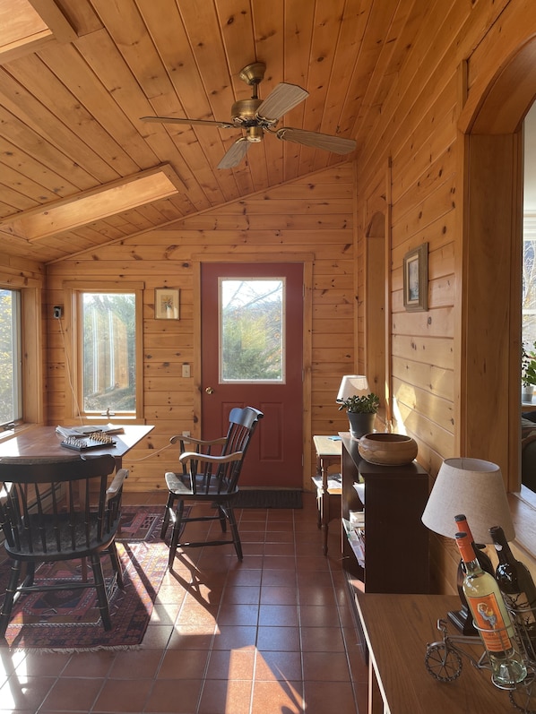 Interior de una cabaña de madera con una mesa de comedor, sillas, un ventilador de techo, una puerta roja y una ventana con vistas a árboles.