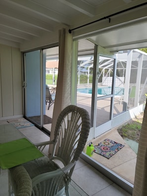 Eine überdachte Terrasse mit einem Korbstuhl, einem Tisch und Blick auf einen Swimmingpool und Gartenmöbel.