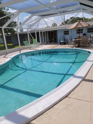 Ein Swimmingpool mit weißem Beckenrand, umgeben von einer überdachten Terrasse mit Stühlen und einem Tisch.