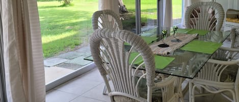 A dining area with wicker chairs and a glass table, large sliding glass doors to a garden, and a ceiling with white beams.