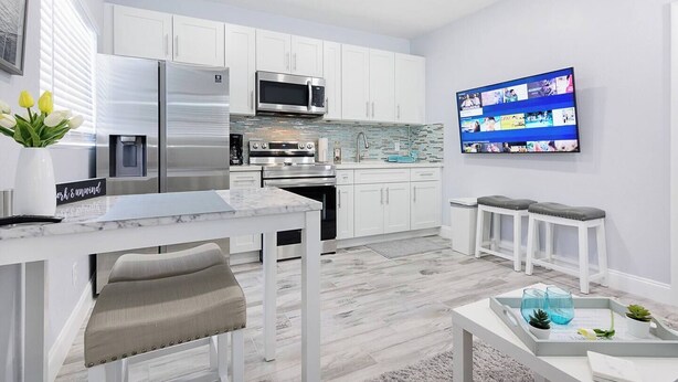 A modern kitchen with white cabinetry, stainless steel appliances, a marble countertop, and a flat-screen TV mounted on the wall. - Welcome to Maya's Blue Lagoon Luxury Suite (Fort Lauderdale)
