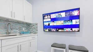 A modern kitchen with white cabinets, a tiled backsplash, and a stainless steel sink. A flat-screen TV mounted on the wall displays various movie posters. - Welcome to Maya's Blue Lagoon Luxury Suite (Fort Lauderdale)