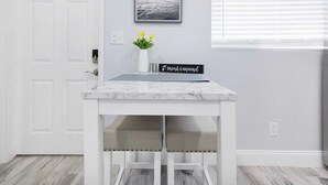 A white table with a marble top, a vase with flowers, and a sign that reads "IT uncork & unwind" in a room with light gray walls, a door, and a window with blinds. - Welcome to Maya's Blue Lagoon Luxury Suite (Fort Lauderdale)