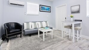 A modern living room with a grey sofa, a black leather chair, a white coffee table, and a white desk with a chair. There are air conditioning units on the wall, a framed picture above the sofa, and a door to the right. - Welcome to Maya's Blue Lagoon Luxury Suite (Fort Lauderdale)