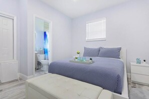 A bedroom with a bed, a bedside table, a nightstand, a window with blinds, a bathroom with a toilet, a shower curtain, and a mirror. - Welcome to Maya's Blue Lagoon Luxury Suite (Fort Lauderdale)