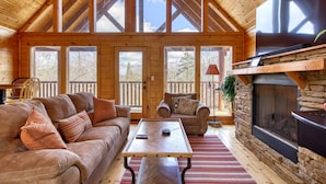 Un acogedor interior de cabaña con techo de madera, ventanas amplias, chimenea de piedra y una zona de estar confortable.
