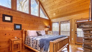 Un dormitorio de cabaña de madera con cama, mesita de noche y ventanas amplias.