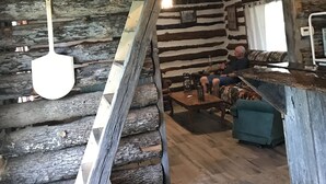 Interior rústico de una cabaña con una escalera de madera, una pared de troncos, una mesa de comedor de madera y un sofá.