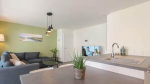 A modern living room with a grey sofa, a small round table, and a potted plant. There is a kitchen area with a sink and a countertop. - Medano 1.2 with balcony by Interhome (Bredene)