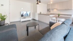 A modern living room with a grey sofa, a small round table, and a TV. The kitchen has white cabinets, a microwave, and a sink. - Medano 1.2 with balcony by Interhome (Bredene)
