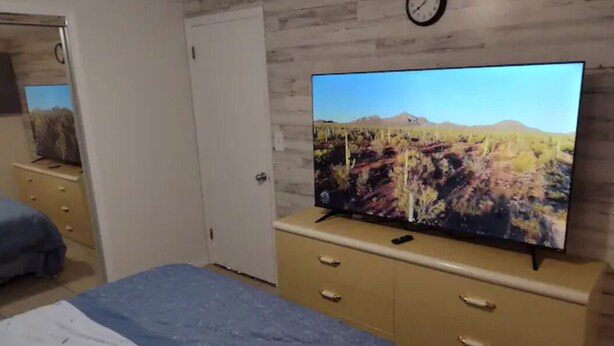 Ein Schlafzimmer mit einem Bett, einem Fernseher auf einer Kommode, einer Uhr an der Wand und einer Tür zu einem anderen Raum.