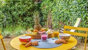 Une table ronde jaune, dressée avec des aliments pour le petit-déjeuner, notamment des fruits, des croissants et des boissons, le tout placé devant un jardin luxuriant.