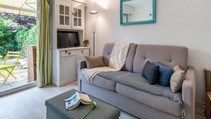 Un salon avec un canapé gris, un téléviseur posé sur un meuble, un miroir au mur et une vue sur la verdure à l’extérieur.