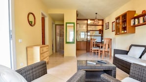 Una sala de estar con muebles de mimbre, una mesa de centro y un sofá. Se observa una cocina al fondo con armarios de madera y una mesa de comedor.