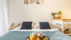 Un lit bien fait, recouvert d’une couverture bleue, avec deux oreillers, un plateau contenant du jus d’orange, une tasse et une tasse rose. On y trouve également une petite table avec une plante et un panier de fruits.