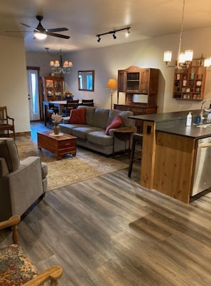 Ein geräumiges Wohnzimmer mit grauem Sofa und Couchtisch aus Holz, eine Küchenzeile mit Holzschränken und einer Arbeitsplatte sowie ein Essbereich mit einer Kronleuchter.