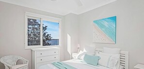 Une chambre à coucher avec un lit, une fenêtre donnant sur un beau paysage, une commode blanche, une chaise et une table de chevet.