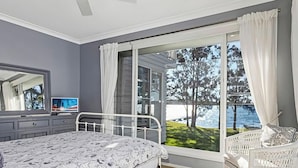 Een slaapkamer met een bed, een televisie, een raam met gordijnen en uitzicht op bomen.