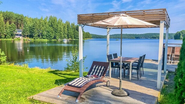 Ein Laube am Seeufer mit Tisch und Stühlen, umgeben von einer Holzterrasse und einer Rasenfläche.
