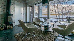 Ein modernes Wohnzimmer mit Kamin, bequemen Sitzmöglichkeiten, einem Couchtisch und einem großen Fenster mit Blick auf eine verschneite Landschaft.