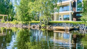 Ein modernes, zweistöckiges Haus mit einem Steg, der in einen ruhigen See führt, umgeben von üppiger Vegetation.