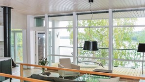 Ein modernes Wohnzimmer mit einem Glastisch, einer Couch und einem Sessel. Ein großes Fenster bietet Blick auf Bäume und ein Gewässer.