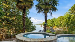 Un jacuzzi rodeado por una piscina de piedra, palmeras y vistas a un lago.