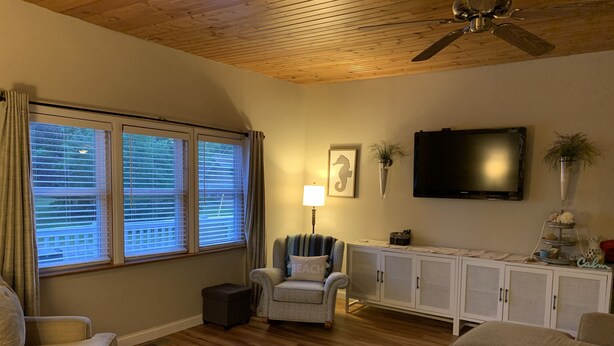 Una sala de estar con techo de madera, un televisor montado en la pared, un mueble blanco y un sillón con reposabrazos.