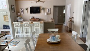Un comedor amplio con una mesa de madera y sillas blancas, un ventilador de techo y un televisor en la pared.