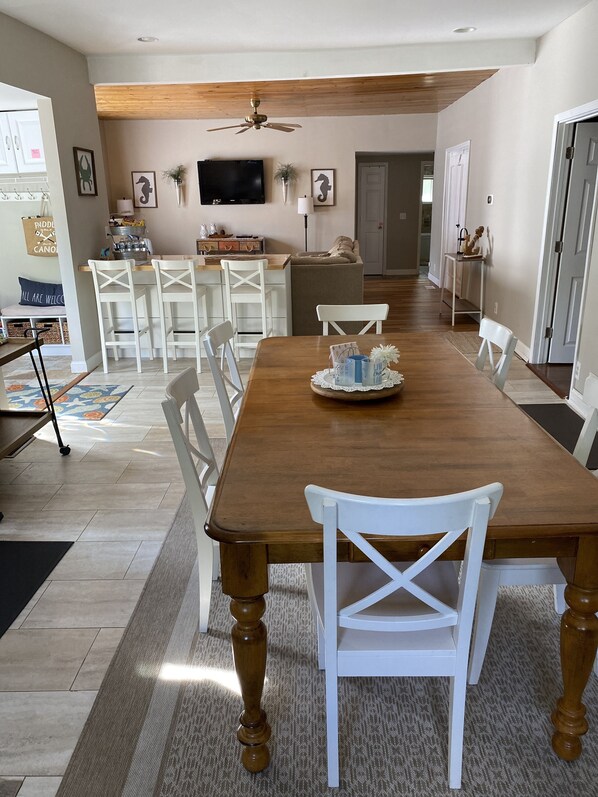 A spacious dining area with a wooden table and white chairs, a ceiling fan, and a television on the wall.