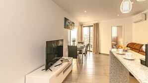 Un salon moderne doté d’un téléviseur à écran plat, d’un coin repas avec une table et des chaises, et d’une cuisine avec un comptoir et des sièges.