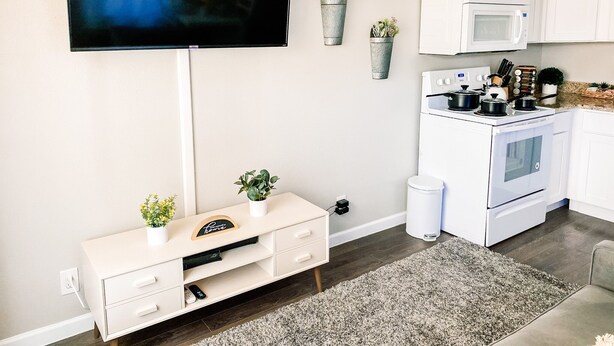 Ett modernt kök med vita skåp, en inbyggd ugn, mikrovågsugn och en spis. En tv är monterad på väggen, och det finns ett litet vitt konsolbord med dekorativa föremål samt en grå matta på golvet.