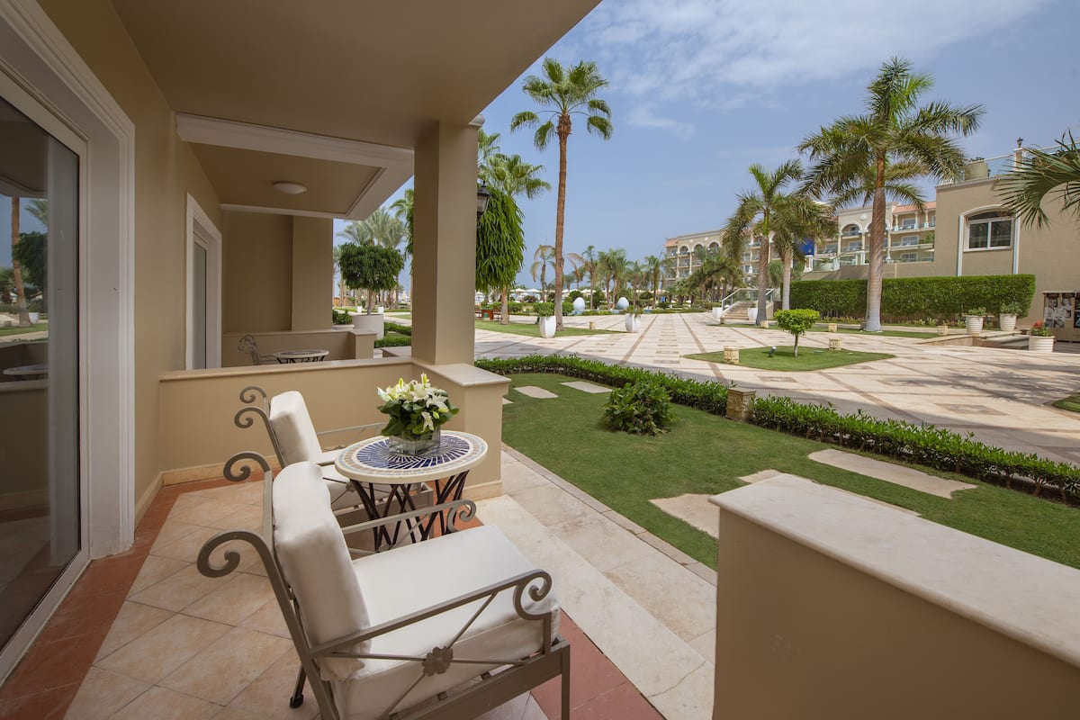 A balcony with a table and chairs, a potted plant, and a view of a well-maintained courtyard with palm trees and a pathway.
