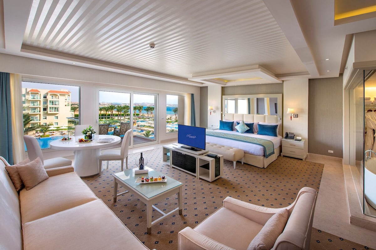 A modern hotel room with a balcony, a TV, a bed with blue bedding, a desk, and a sofa.