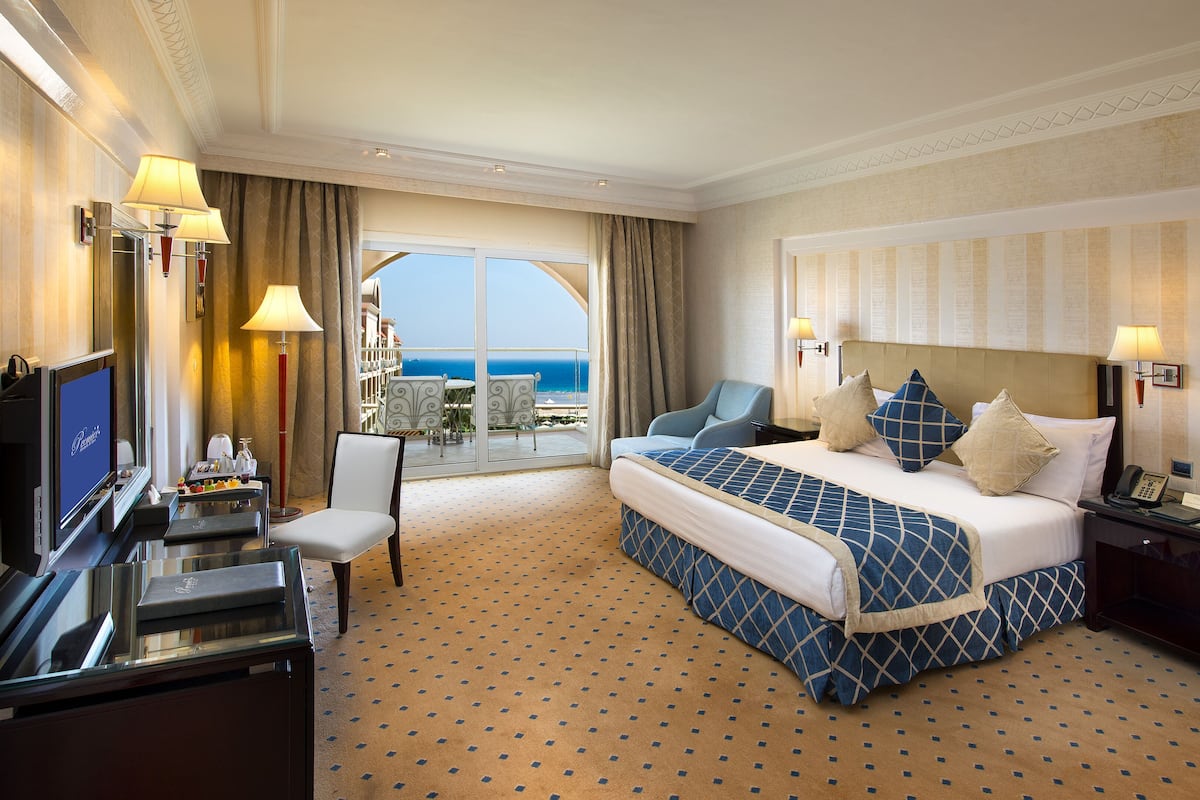 A hotel room with a large bed, a desk with a TV, a chair, and a balcony with a view of the ocean.