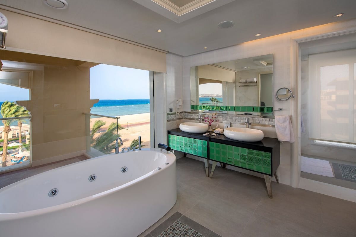A modern bathroom with a large bathtub, dual sinks, and a view of the beach.