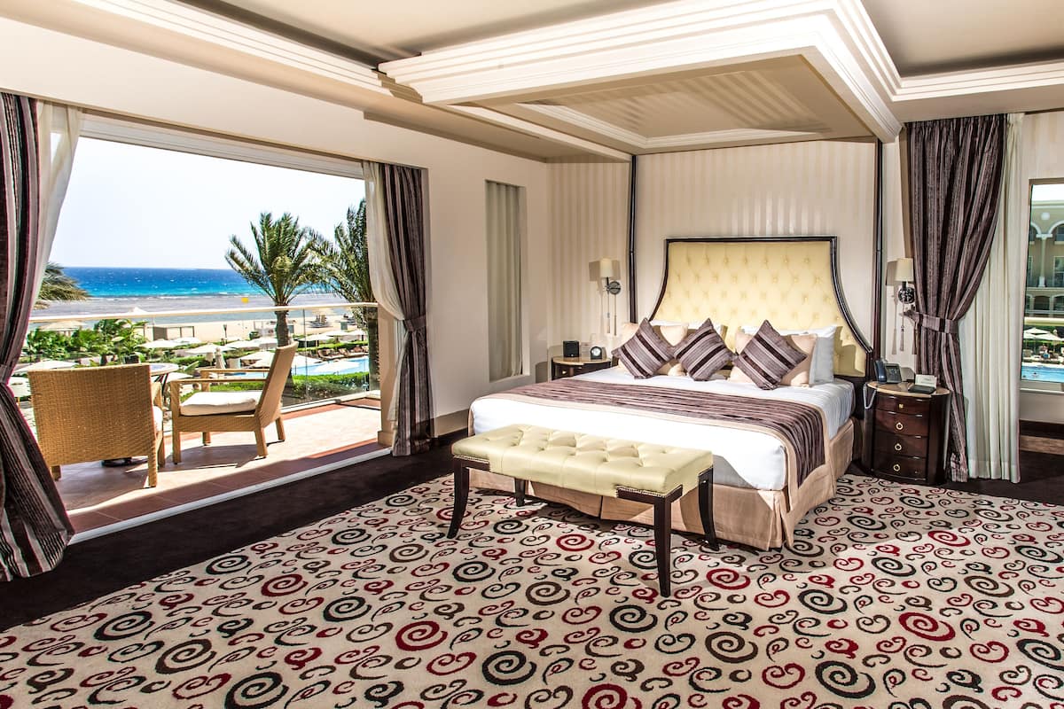 A spacious hotel room with a large bed, a view of the pool and beach, and a balcony with wicker furniture.