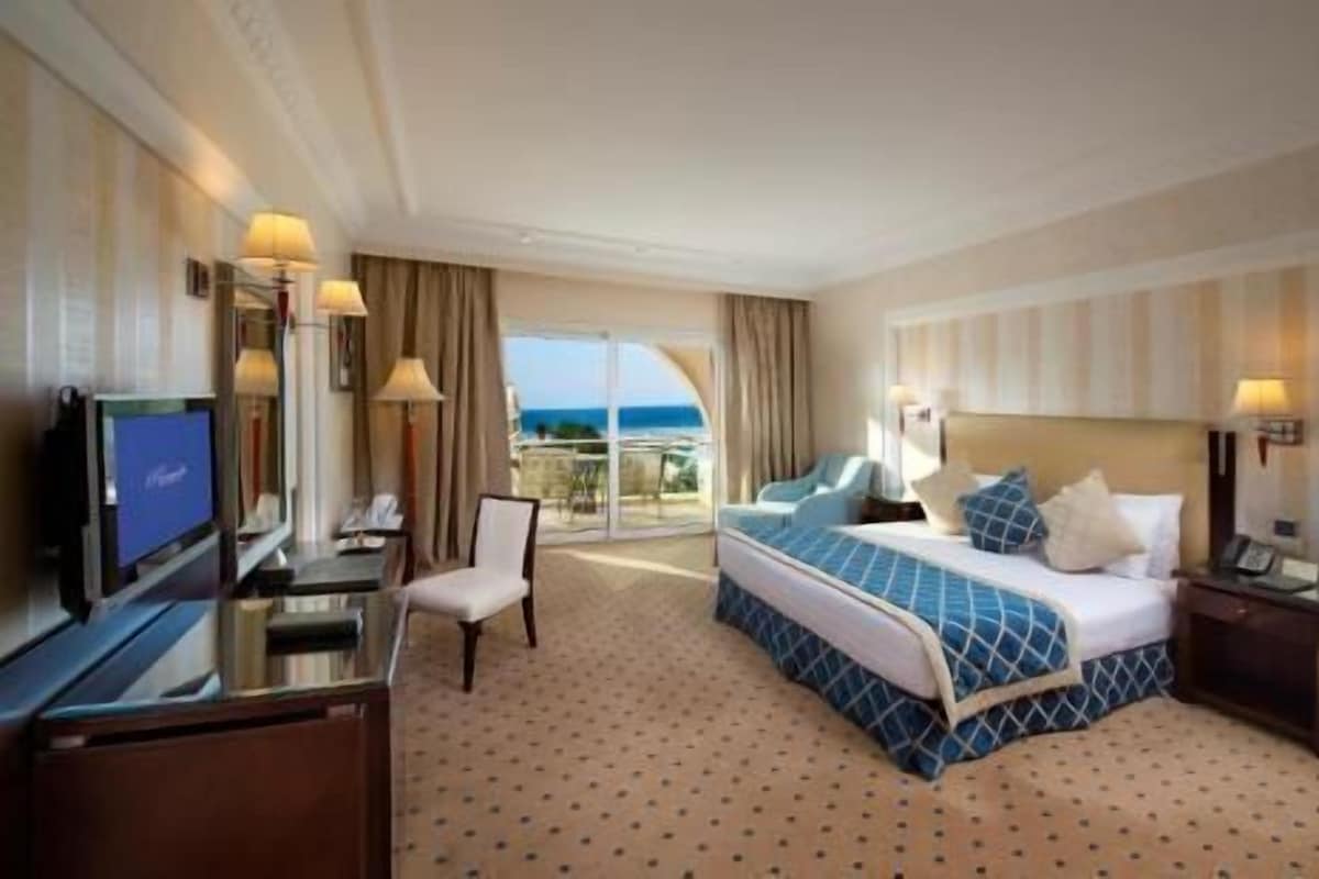 A hotel room with a bed, a desk, a chair, a TV, and a view of the ocean.