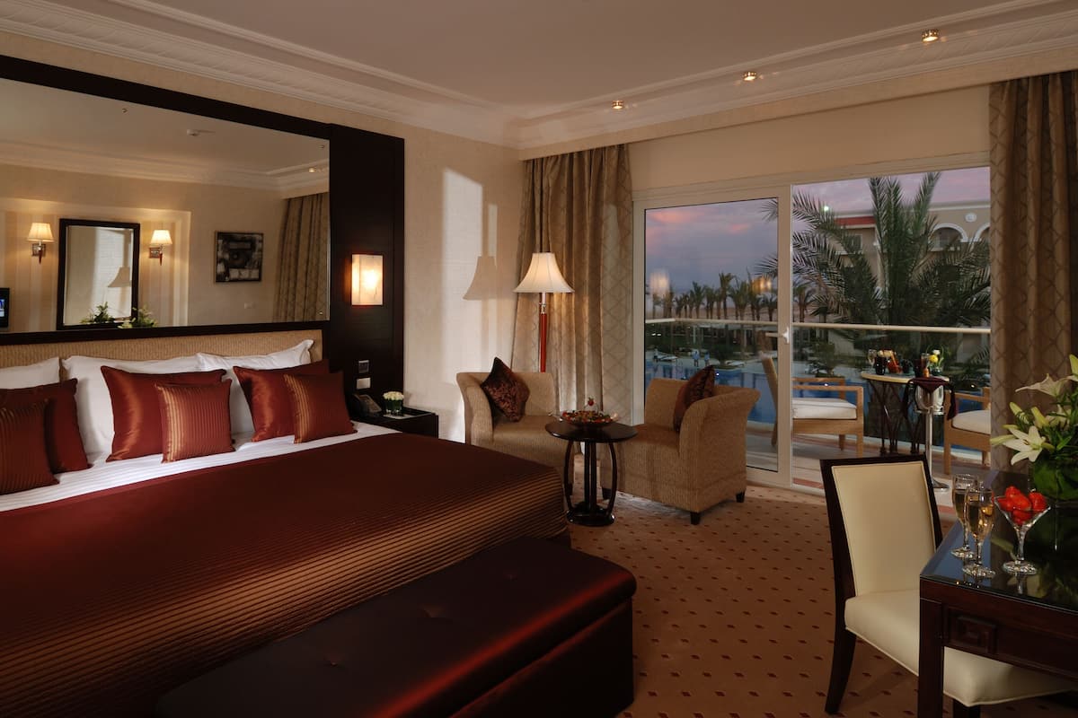 A hotel room with a large bed, a sofa, a small table with a vase, and a view of a pool and palm trees.