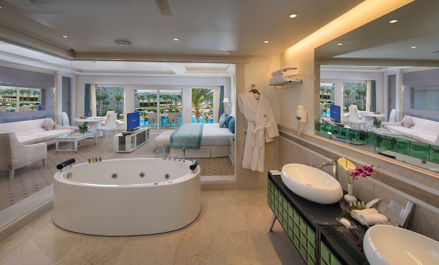 A modern bathroom with a large bathtub, a vanity with a large mirror, and a view of a pool and palm trees.