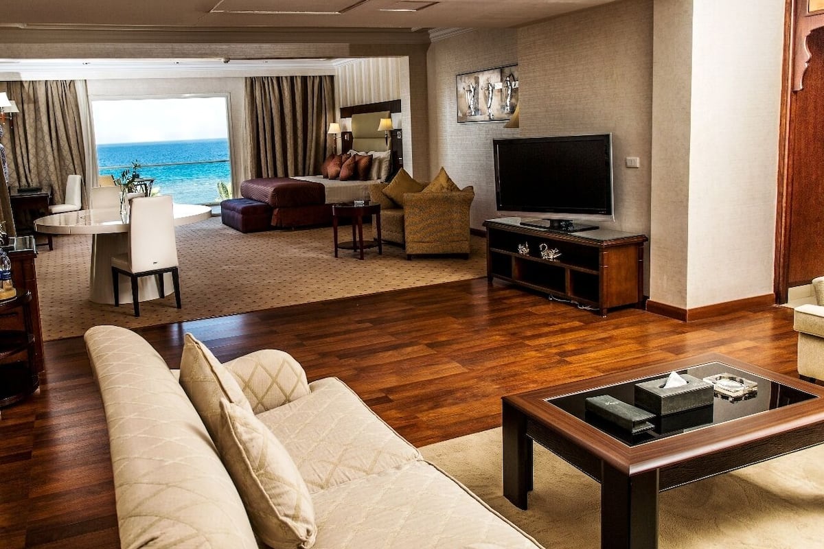 A spacious living room with a large sofa, a coffee table, a TV, and a view of the ocean.
